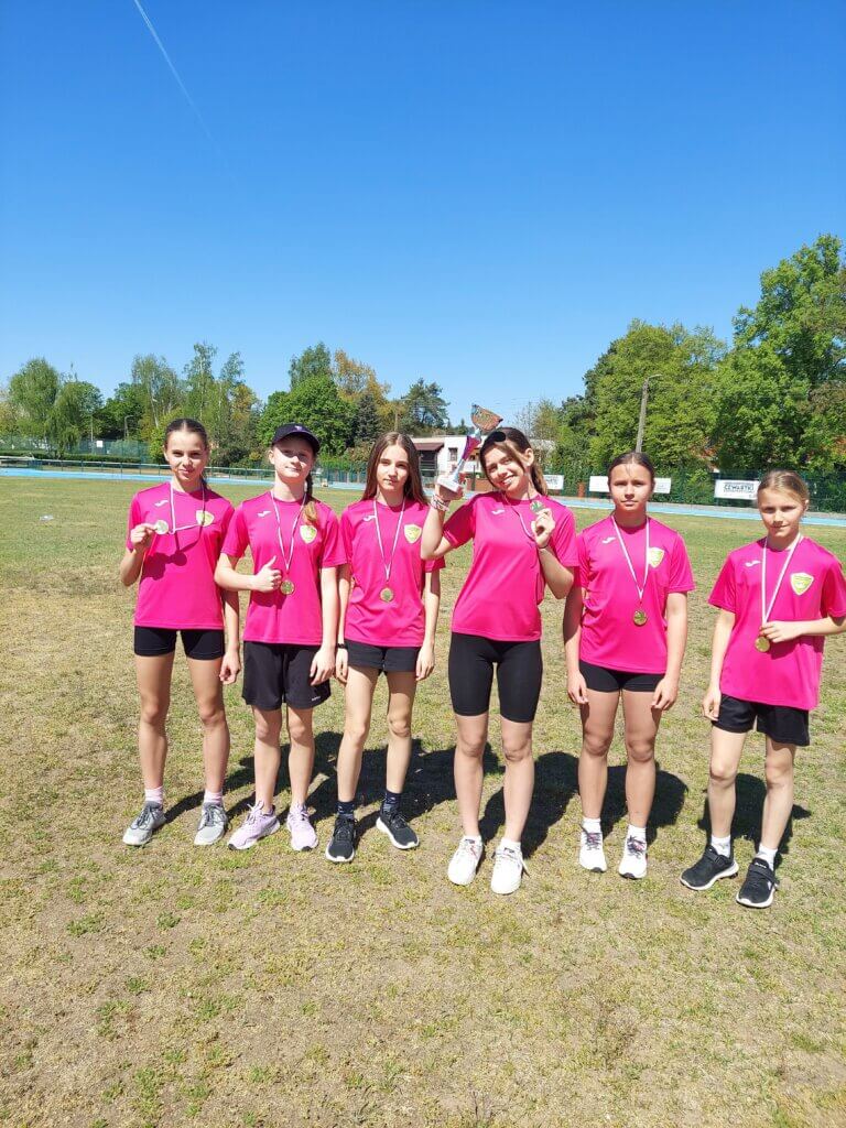 Sześć dziewcząt w różowych koszulkach sportowych stoi na boisku trzymając medale i puchar. Wszystkie są uśmiechnięte i pozują po zwycięstwie w zawodach lekkoatletycznych. W tle widać zielone drzewa, bieżnię oraz niebieskie niebo.